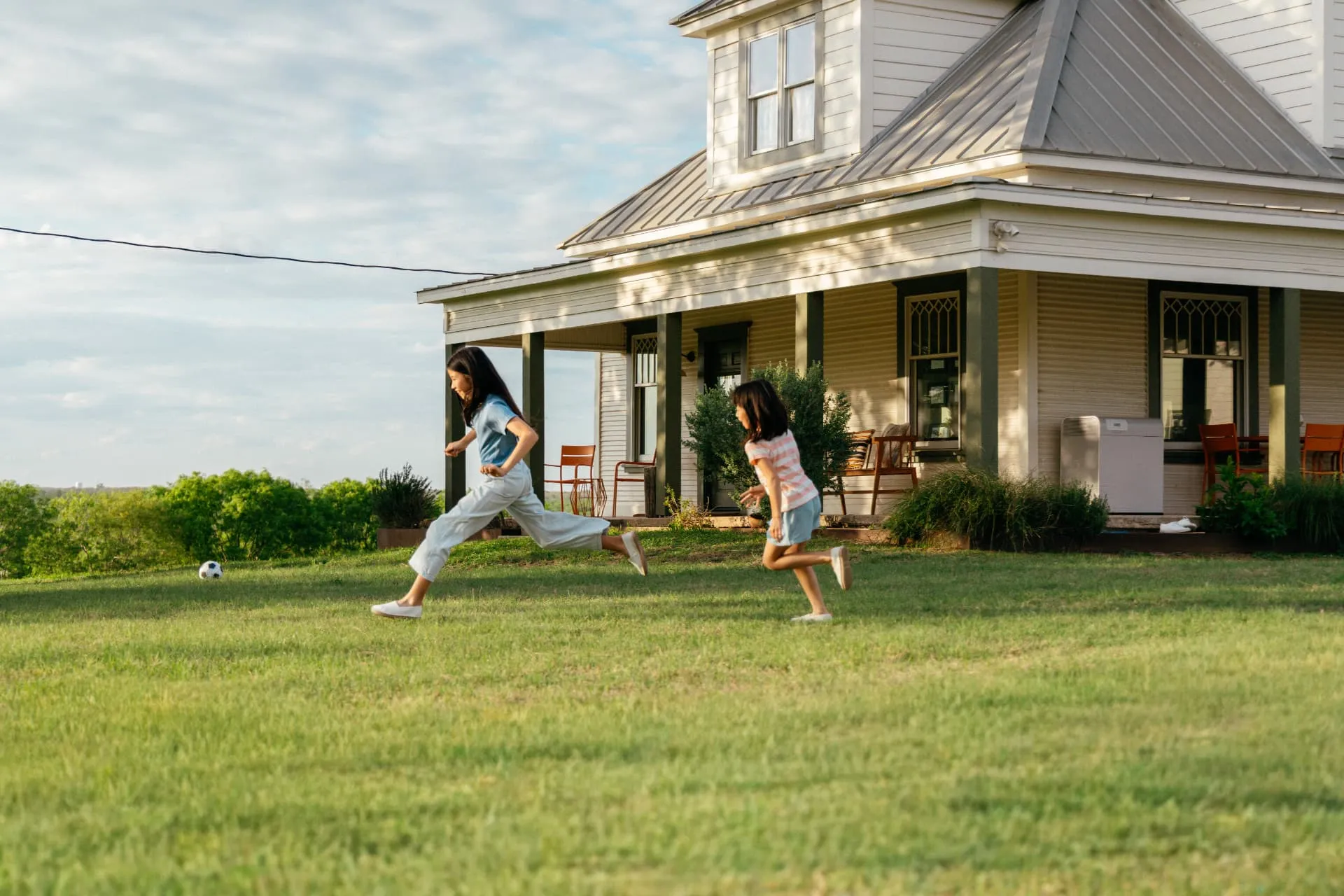 Kids playing soccer in the yard of a Texas farmhouse with a Base Power battery on the porch