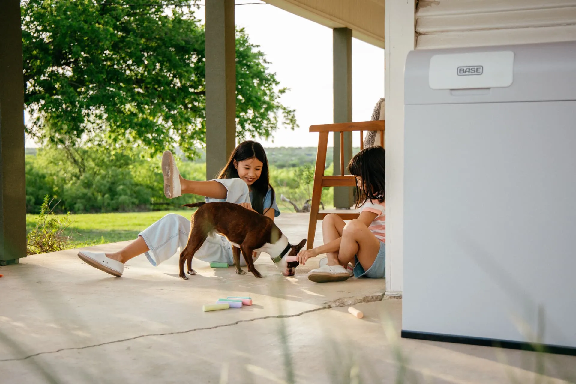 Two kids drawing with chalk and playing with a dog on a porch next to a Base Power battery